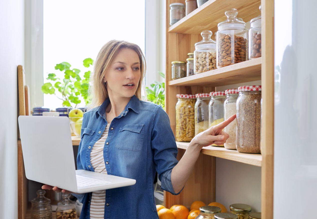 Woman in home kitchen pantry, holding laptop, food blog, online nutritionist consultation